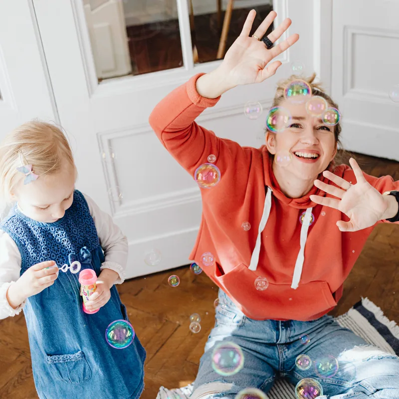 A young sitter kneels on the floor of a bright room, laughing with a toddler in dungarees as they chase soap bubbles through the air.