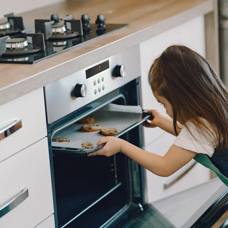 A girl slides a baking tray of cookies into the oven in a bright modern kitchen.