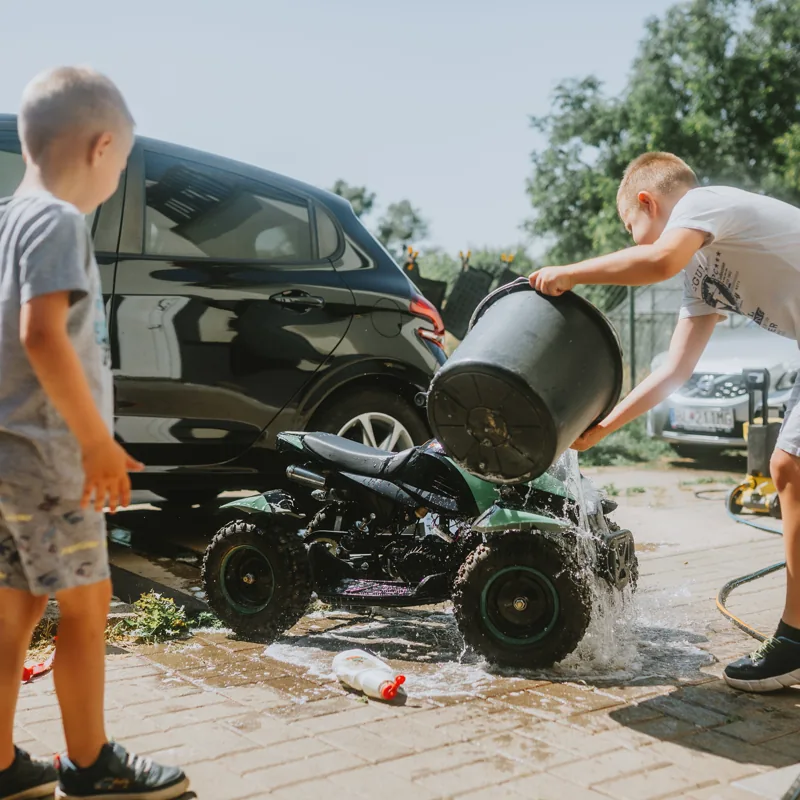 Two boys in shorts and t-shirts wash a small vehicle in a sunny driveway, tipping a bucket of soapy water over it.