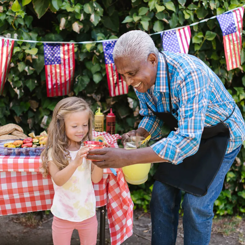 A girl is handed a cup of lemonade across an outdoor table decorated with bunting on a bright summer day.