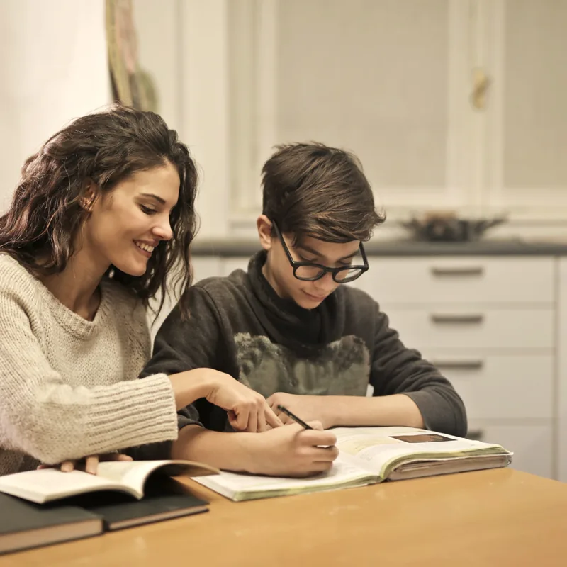 A cheerful young woman sits beside a boy at a wooden table, pointing at his notebook while he works through homework.